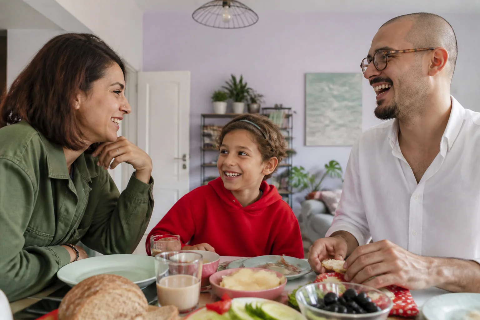 Bir aile akşam yemeği masasında birlikte gülerek sohbet ediyor, ortada kırmızı kazaklı bir kız çocuğu