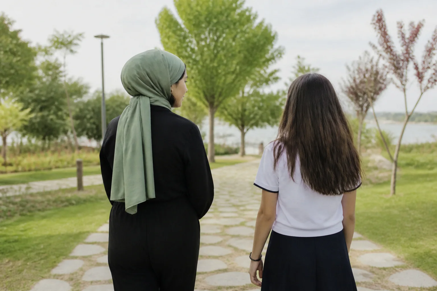 Ağaçlıklı bir yolda yan yana yürüyen başörtülü bir anne ile ergen kızı, arkadan görünüyor
