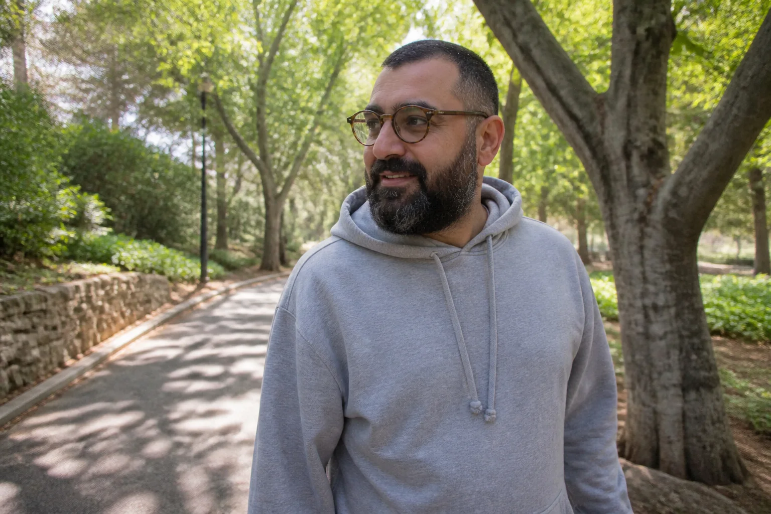 Ağaçlıklı bir parkta gri kapüşonlu sweatshirt giymiş gözlüklü adam yürürken gülümsüyor